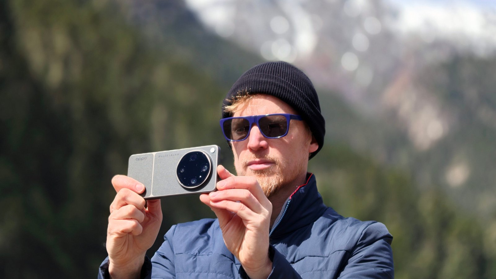 Man in beanie hat and sunglasses taking a picture with the Oppo Find X9 Ultra, there are snowy mountains behind him